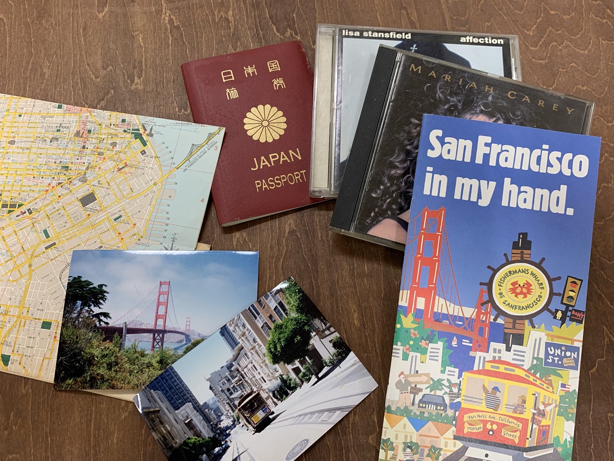 サンフランシスコ旅行を思わせる平置き写真。日本のパスポーツ、地図、ケーブルカーとゴールデンゲートブリッジの写真、マライア・キャリーとリサ・スタンスフィールドのCD、旅行パンプレットなどが木のテーブルに並ぶ。