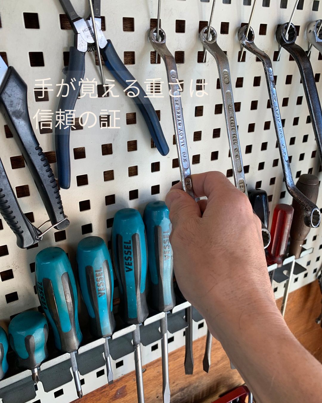 壁に整然と掛けられた工具棚から、手がメガネレンチを取り上げる瞬間。金属の重さと使い込まれた質感が伝わる。
